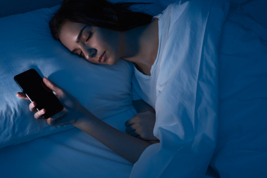 Imagem de uma mulher dormindo em sua cama com celular na mão mostrando causas de má produção de Melatonina
